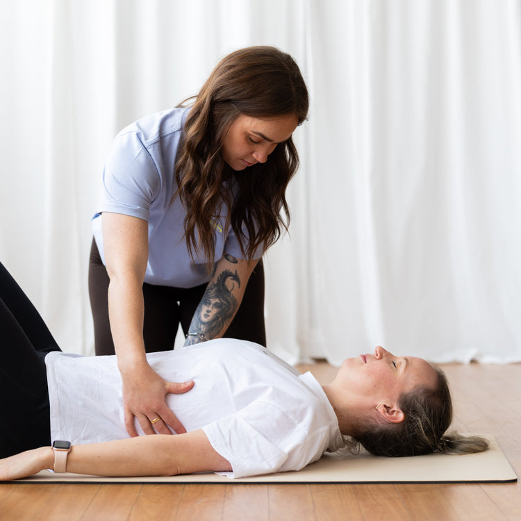 A woman in a white shirt and black pants lies on a yoga mat while another woman in a light blue shirt and black pants leans over her, demonstrating a massage or stretching technique in a bright room with white curtains and a wooden floor.